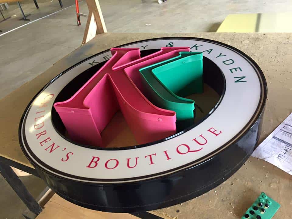 A round store sign with a black frame and white background features the text "Keely & Kayden Children's Boutique" in pink and green capital letters. The sign includes a large "K" in pink and green 3D letters in the center. It's propped up on a wooden table, showcasing top-quality Custom Sign Services Orange County can offer.
