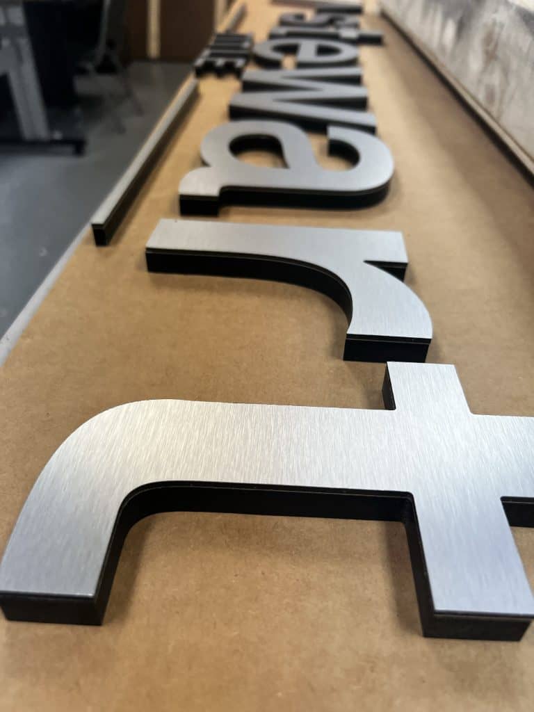 Close-up view of brushed metal letters on a flat surface, laid out to form a sign by a Sign Company in Orange County. The background shows part of a workshop with tools and materials scattered. The letters are in a modern, bold typeface.