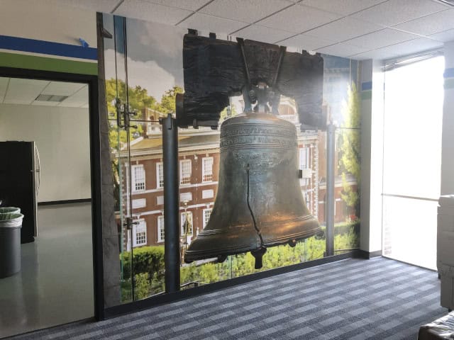 The office interior showcases a large mural of the Liberty Bell with Independence Hall in the background. Carpeted floors, overhead lights, and a window on the right side let in natural light, while custom signs add a personalized touch to this inspiring workspace.