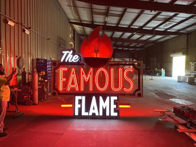 Inside a spacious warehouse, a large custom sign reading "The Famous Flame" glows brilliantly. Featuring a red flame with a fork design above the text, this striking piece of custom signage casts its vibrant glow throughout the dimly lit space.