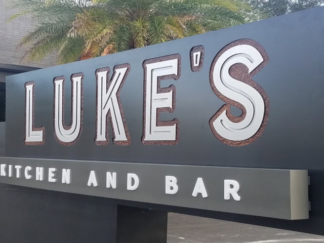A custom sign for "Luke's Kitchen and Bar" features large, bold white letters with a textured border. The background showcases palm fronds and part of a building, exemplifying top-notch Business Signs in Orange County.