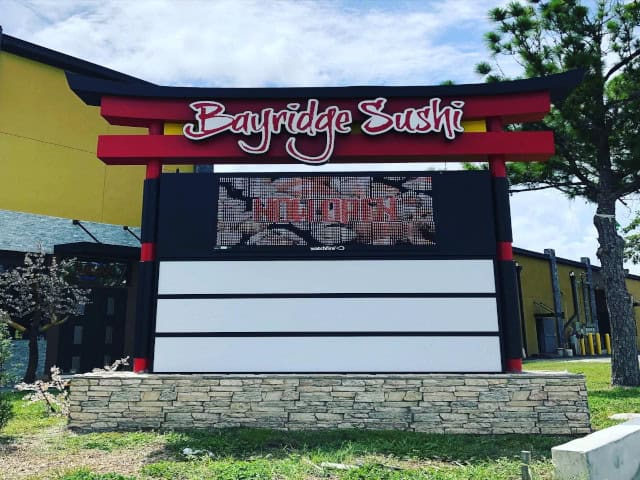 Sign for "Bayridge Sushi" with red and black accents and a stone base, located in front of a building. The sign features a digital display showing "Welcome". There is a tree and some greenery surrounding the sign, typical of high-quality work by Business Signs Orange County, with part of the building visible in the background.