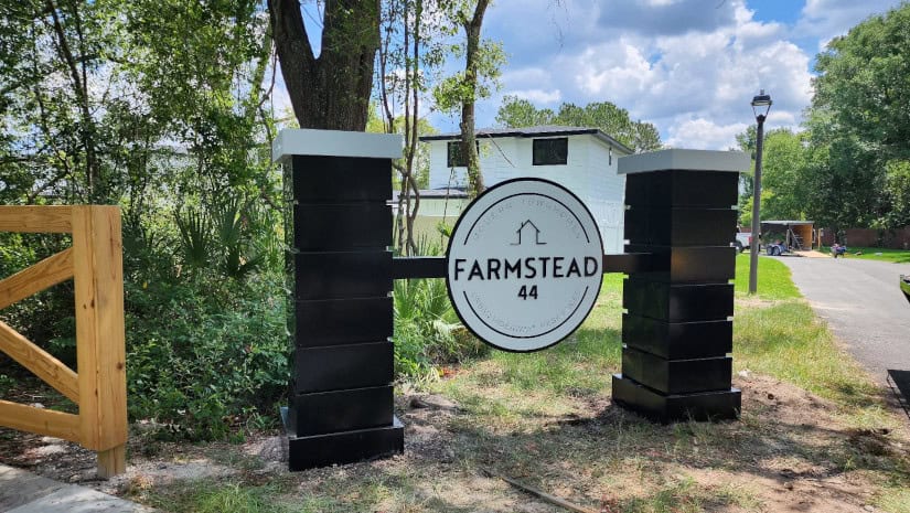 A sign reading "Farmstead 44" is mounted between two black pillars in a rural setting. Trees and greenery are seen in the background, along with part of a white building and a paved path. Expertly crafted by a Sign Company in Orange County, the sign stands against a bright sky with scattered clouds.