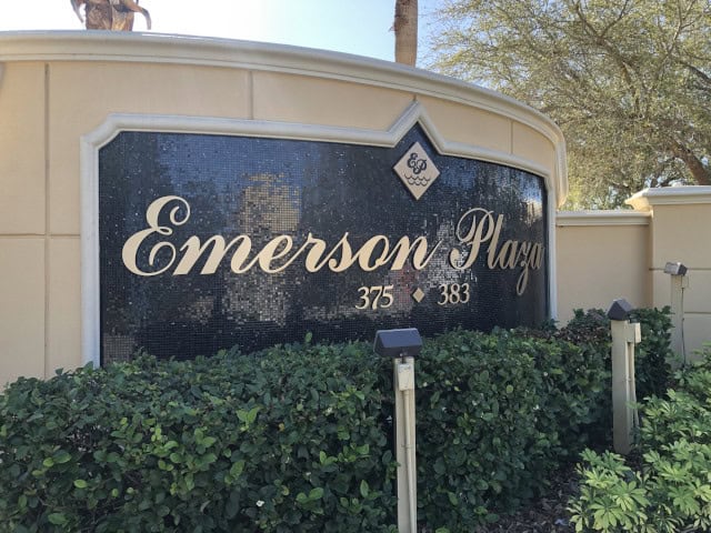 A sign reads "Emerson Plaza" in elegant script, mounted on a curved wall adorned with black tiles and flanked by greenery. The numbers "375" and "383" are listed below the text. Created by a leading Sign Company in Orange County, trees and part of a statue are visible in the background.
