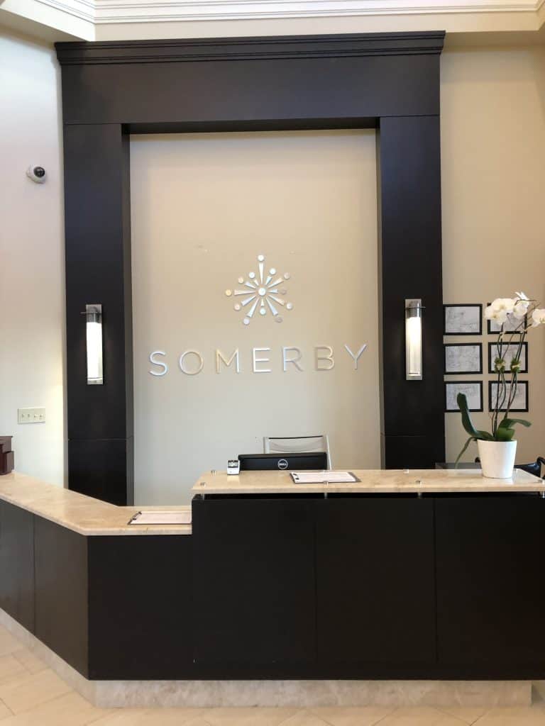 A reception desk with a marble countertop and dark wood paneling stands proudly. Behind it, a logo featuring an abstract starburst design and the name "SOMERBY" is showcased, courtesy of Custom Sign Services Orange County. The area is illuminated by two wall sconces and a potted orchid adds an elegant touch.