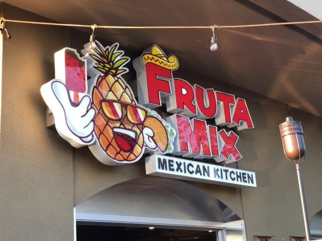 A colorful sign above a storefront reads "Fruta Mix Mexican Kitchen." The sign, possibly crafted by custom sign services in Orange County, features a cartoon pineapple character wearing sunglasses and making a thumbs-up gesture, with additional images such as a sombrero and a palm tree in the design.