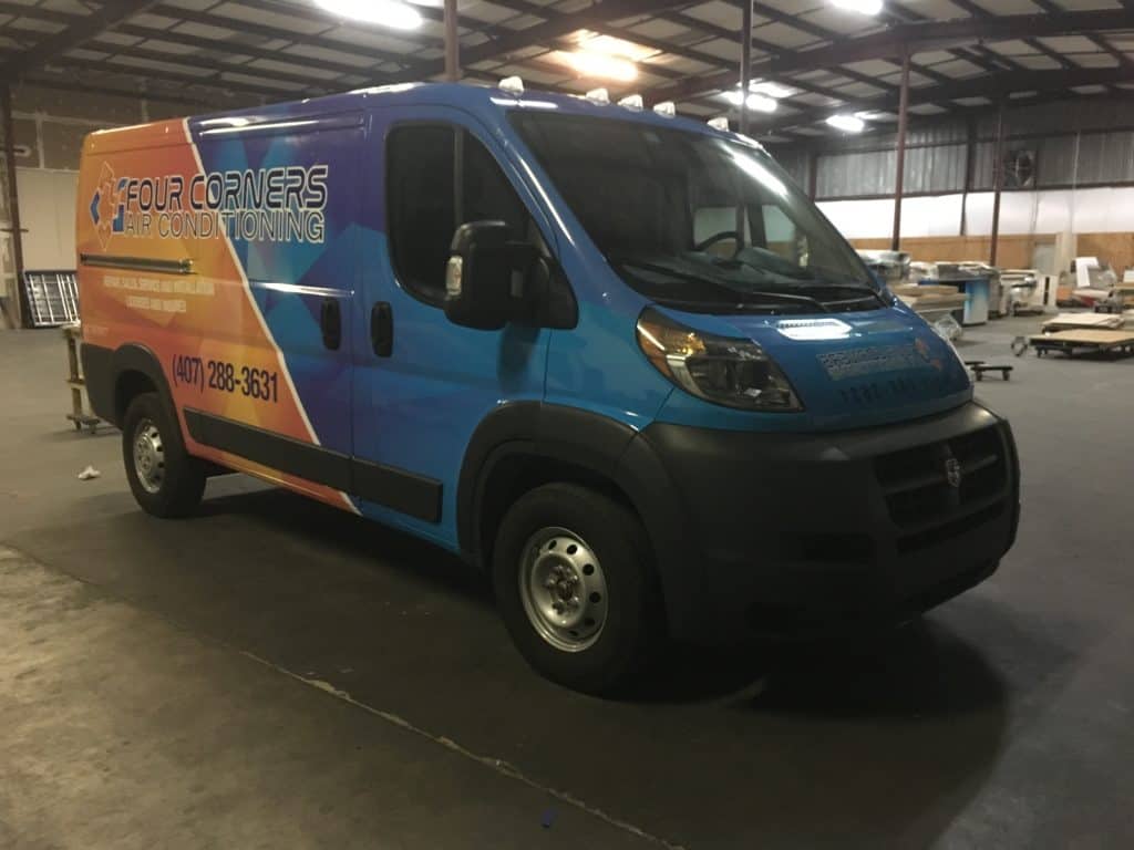 A blue and orange van with "Four Corners Air Conditioning" and a phone number printed on the side is parked inside a large warehouse. The van, proudly showcasing branding from a renowned Sign Company in Orange County, features air conditioning services, while the warehouse contains various equipment and supplies.