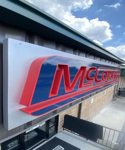 A sign for "McCorvey" in large red and blue letters is mounted on the side of a building. The building, featuring a metal exterior with a brick facade below the sign, showcases the quality of Custom Sign Services Orange County. The sky above is partly cloudy.