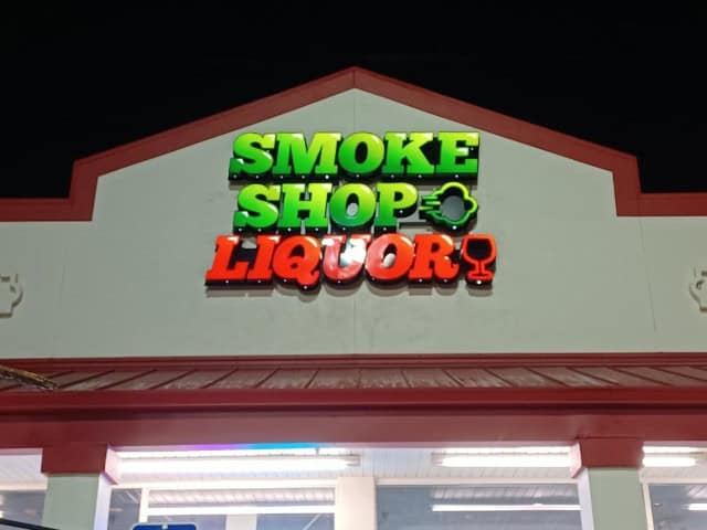 A store sign in bright colors on a building reads "Smoke Shop Liquor" with "Smoke Shop" in green and "Liquor" in red, accompanied by an image of a liquor glass. The building, crafted by Custom Sign Services Orange County, has a white exterior with red trim. The photo is taken at night.