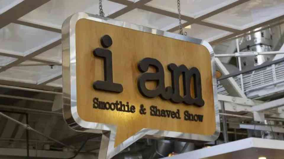 A wooden sign in the shape of a speech bubble hangs from the ceiling, displaying the text "i am" in large letters, and "Smoothie & Shaved Snow" in smaller letters underneath. The background shows the interior structure of a building with exposed beams, showcasing quality work by Business Signs Orange County.