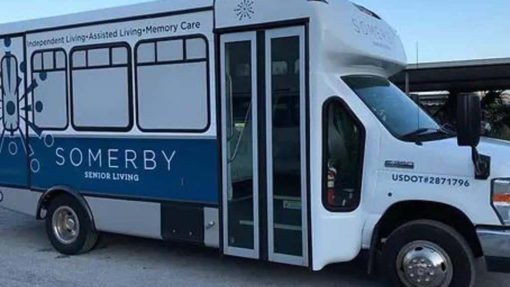 A bus with the branding "Somerby Senior Living" is parked, featuring signage for independent living, assisted living, and memory care. The bus has a blue and white color scheme and displays the USDOT number 2871796 on the side. Expertly crafted by Custom Sign Services Orange County.