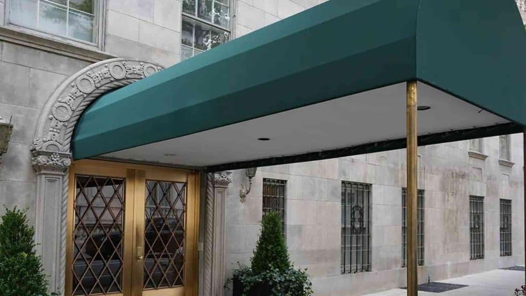A building entrance featuring a green fabric awning supported by gold-colored poles. The entrance has double, diamond-patterned glass doors with gold frames and is flanked by potted greenery. The facade, crafted from stone with arched decorative elements above the doors, showcases the quality of Awnings Orange County.