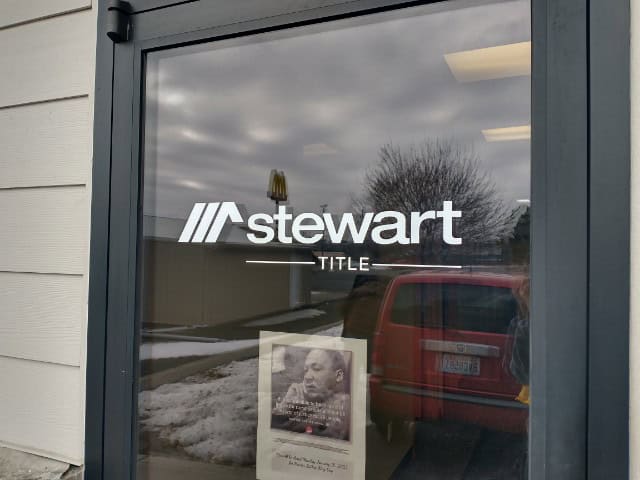 A glass door displaying the "Stewart Title" logo in white font. Behind the door, a red car, snow on the ground, and an overcast sky are visible. A small poster with a black-and-white photo and text is taped to the bottom of the door—courtesy of Custom Sign Services Orange County.