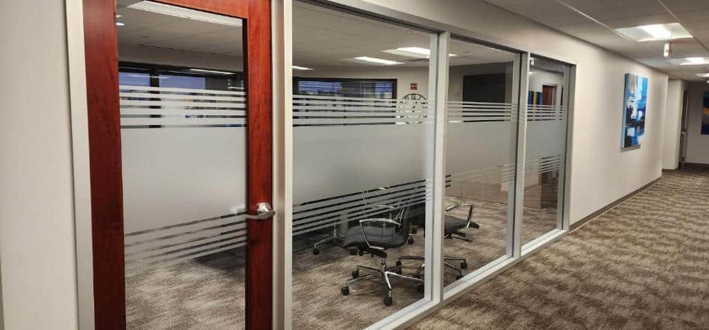 A modern office hallway with a glass-walled conference room on the left, featuring frosted stripes for privacy and filled with office chairs. The carpeted hallway includes a few closed doors, a framed picture on the walls, and Business Signs Orange County for clear navigation.