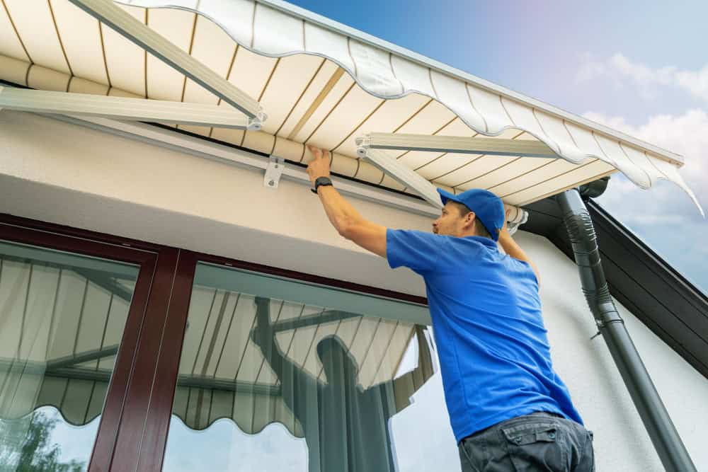 A person in a blue shirt and cap installs a retractable awning on the exterior of a house, positioned above large windows reflecting the sky. The awning is white with beige stripes, offering shade much like the quality installations provided by Sign Company Orange County. The sky is blue with some clouds.