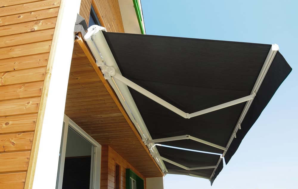 A retractable, dark-colored awning from Awnings Orange County is extended over a wooden building's exterior, providing shade. The awning has a sturdy, white metal frame and is positioned above a doorway, with clear blue skies in the background.