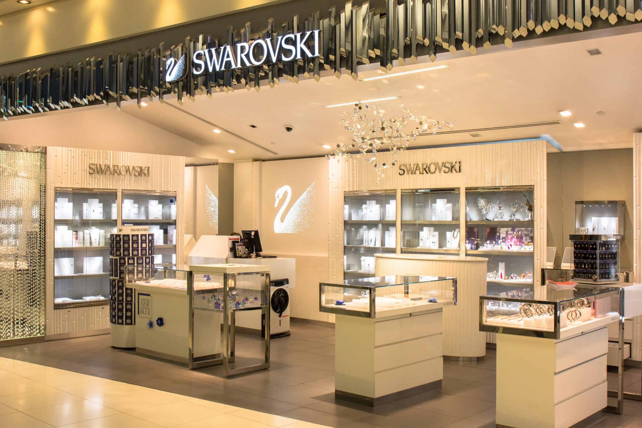 A Swarovski store featuring a modern interior with illuminated display cases presenting various crystal products. The store, enhanced by a chandelier and eye-catching business signs from Orange County, proudly showcases the Swarovski logo and swan emblem, creating an elegant shopping environment.
