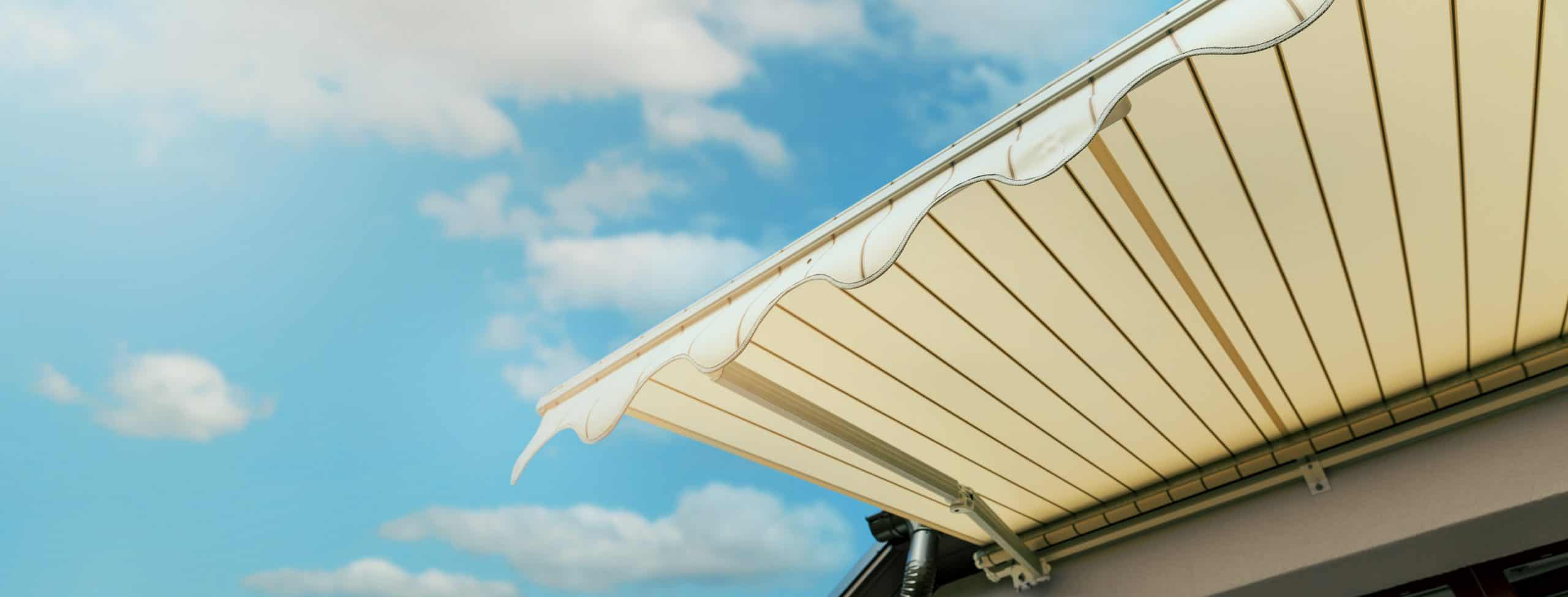 A beige retractable awning extends over a balcony, providing shade under a bright blue sky with scattered fluffy clouds. The awning features wavy scalloped edges, adding a decorative touch. Perfect for Awnings Orange County and accentuating the serene sky above, this setup highlights both style and functionality.