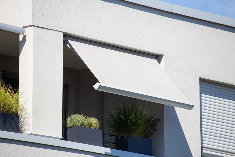 A close-up of a modern building's balcony featuring a beige retractable awning, provided by a Sign Company in Orange County. Below the awning are several potted plants, including grasses and shrubs in square containers. The balcony's clean lines contrast with the rolled-up white shutter next to it.