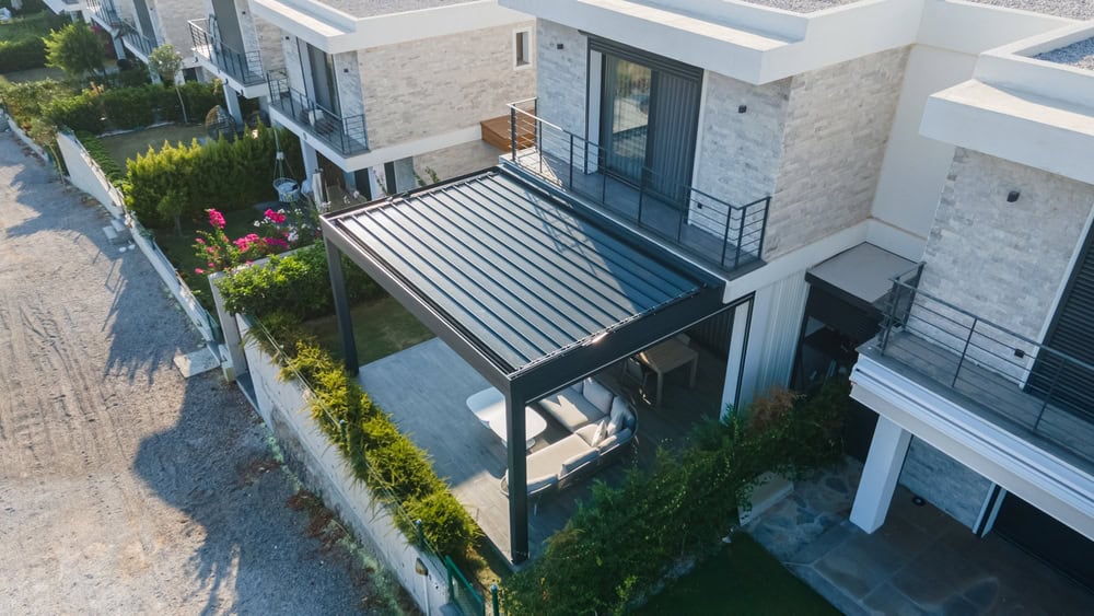 Aerial view of a modern two-story house with a large patio covered by a metal pergola. The ground level features an outdoor seating area surrounded by greenery, and the house exterior is made of light stone with balconies on the upper floors, much like the elegant designs admired in Sign Company Orange County projects.